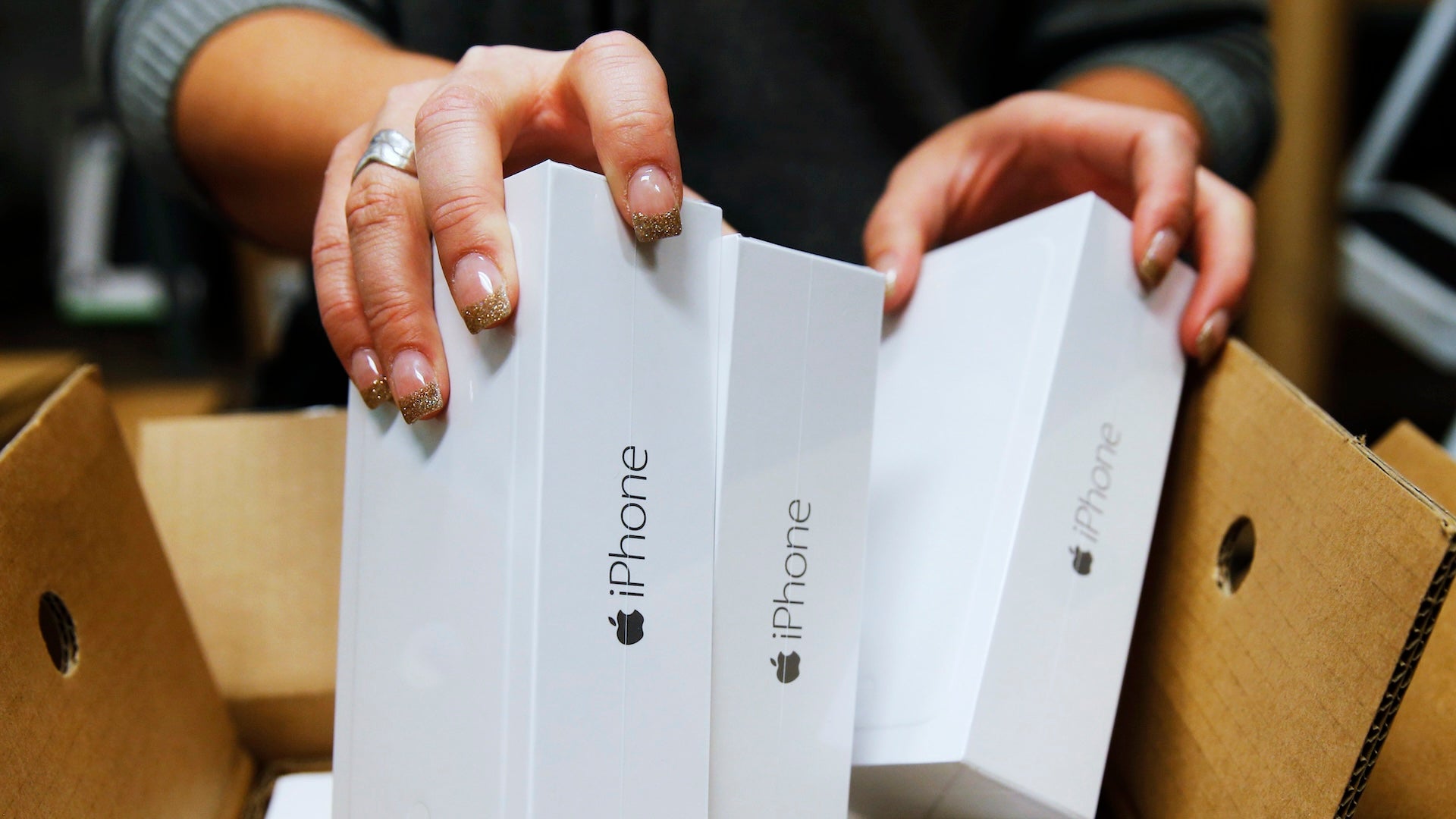 Several iPhones in boxes are lifted by hands from a larger box.