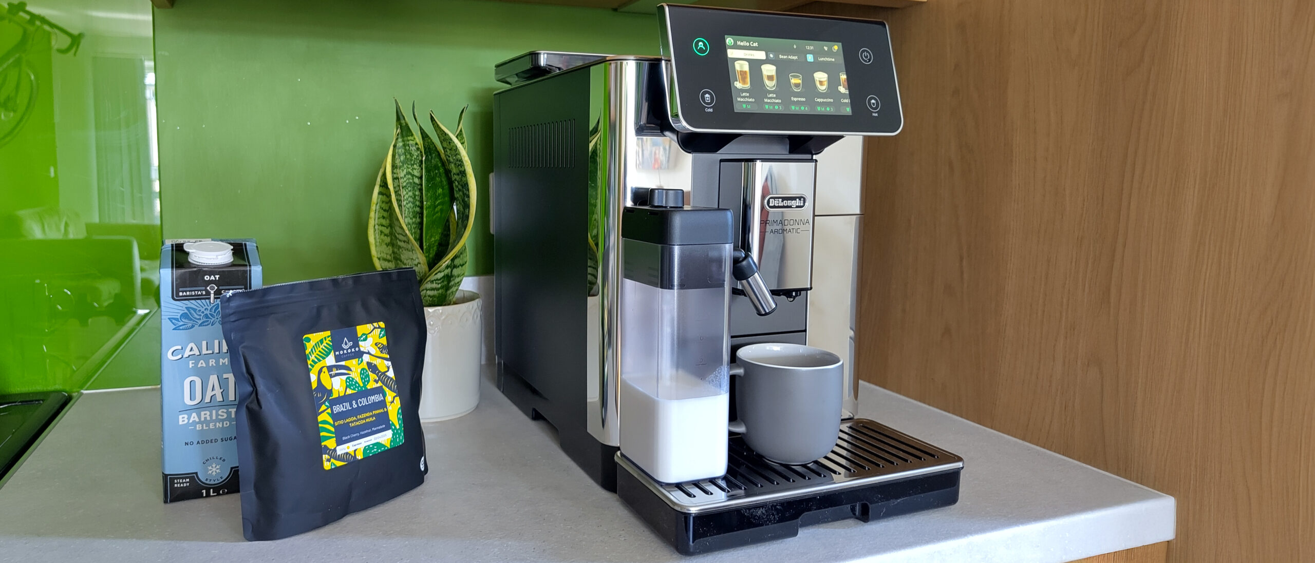 De'Longhi Primadonna Aromatic espresso machine on kitchen counter with coffee beans, oat milk, and snake plant