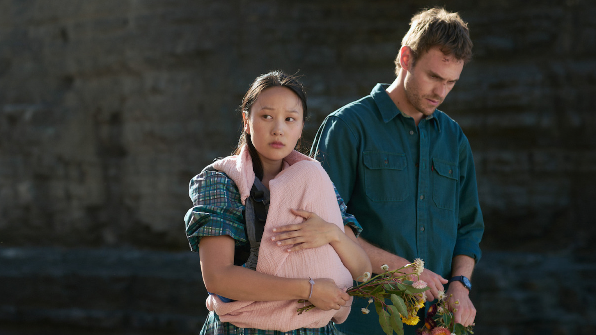 A still from the Netflix series The Survivors showing a man and a woman on a beach with a cliff behind them. The woman is carrying a baby in a pink blanket, although the baby is not visible.