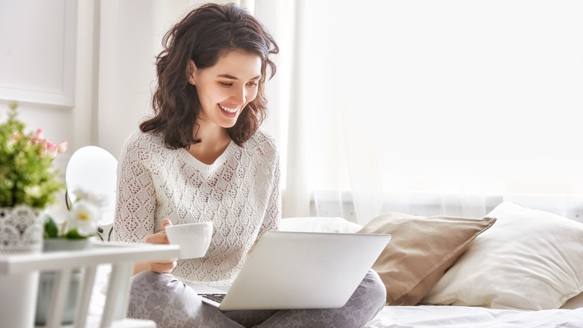 Happy woman sitting on a bed with a coffee and a Windows 11 laptop